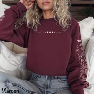 Pode incluir: Uma camisola de manga comprida cor de vinho com um gráfico branco das fases da lua centrado no peito. As mangas estão decoradas com uma impressão botânica branca detalhada de flores silvestres, fetos e traças ao longo dos braços.