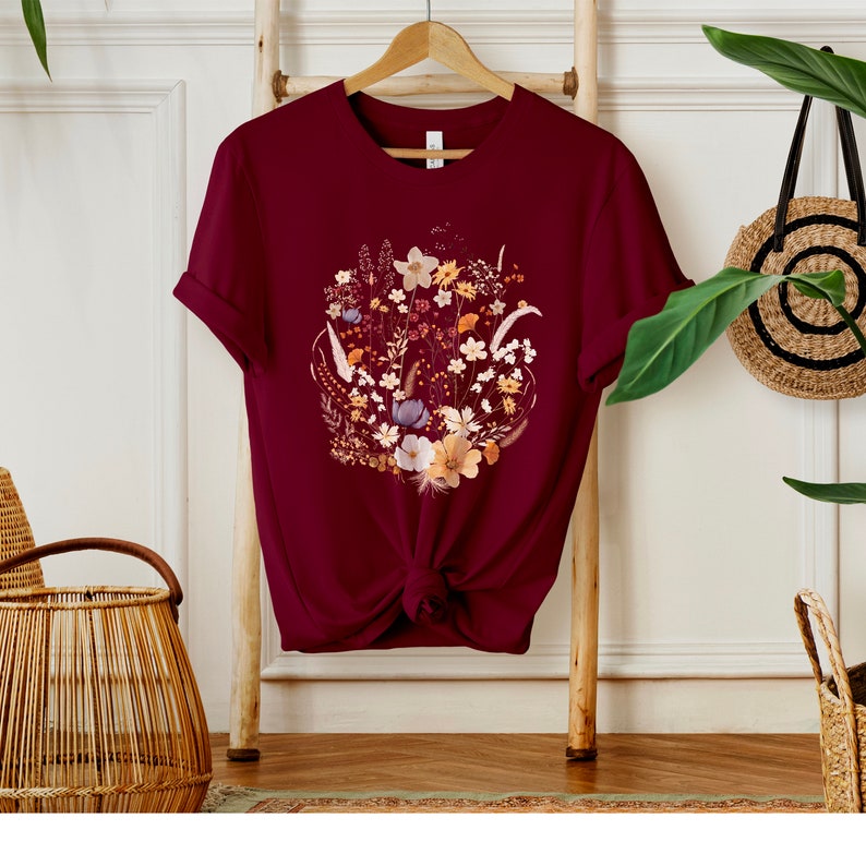 May include: A maroon t-shirt with a floral print featuring a variety of flowers in shades of brown, yellow, white, and blue.