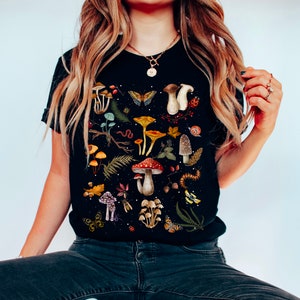 May include: Black t-shirt with a colorful mushroom and forest illustration. The illustration includes mushrooms, butterflies, leaves, acorns, and other forest elements.