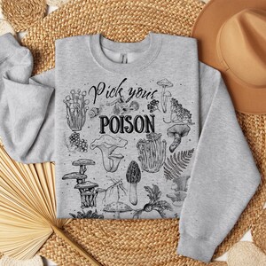May include: A light grey sweatshirt with a crew neck featuring a black and white illustration of various mushrooms and the text "Pick your POISON". The sweatshirt is made of a soft material.