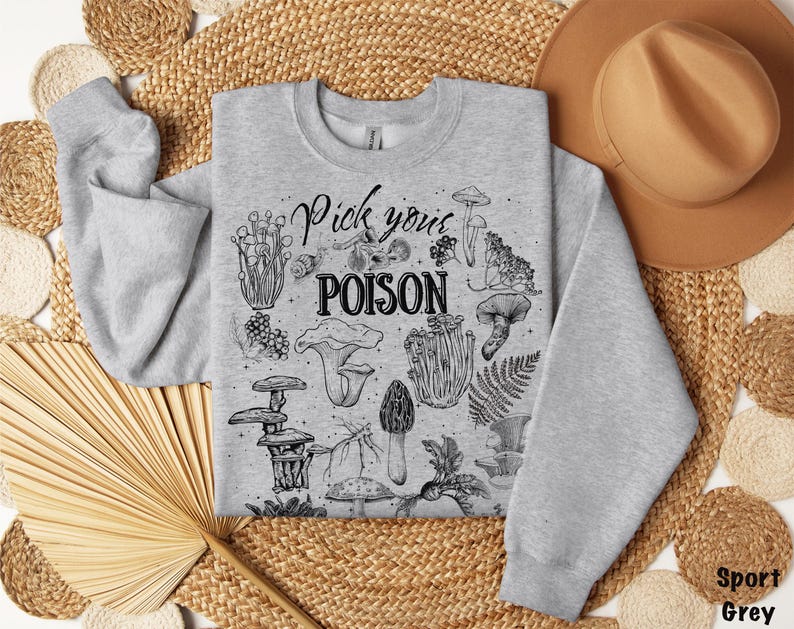 May include: A light grey sweatshirt with a crew neck featuring a black and white illustration of various mushrooms and the text "Pick your POISON". The sweatshirt is made of a soft material.