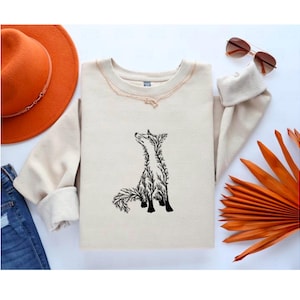 May include: A beige crewneck sweatshirt featuring a black graphic illustration of a fox composed of intricate tree branch patterns. The sweatshirt is displayed alongside an orange wide-brimmed hat, blue denim jeans, brown sunglasses, and a dried orange palm leaf.