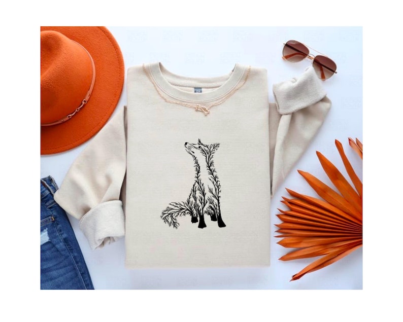 May include: A beige crewneck sweatshirt featuring a black graphic illustration of a fox composed of intricate tree branch patterns. The sweatshirt is displayed alongside an orange wide-brimmed hat, blue denim jeans, brown sunglasses, and a dried orange palm leaf.