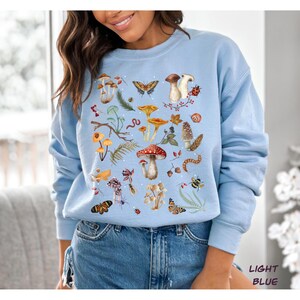 K&ouml;nnte beinhalten: Ein hellblaues Sweatshirt mit einer bunten Illustration von Pilzen, Schmetterlingen und anderen Waldelementen. Das Design zeigt eine Vielzahl von Pilzen in verschiedenen Formen und Gr&ouml;&szlig;en sowie Schmetterlinge, Marienk&auml;fer und andere Insekten. Das Sweatshirt hat einen Rundhalsausschnitt und lange &Auml;rmel.