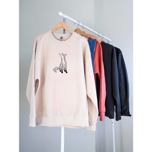 May include: A beige crewneck sweatshirt featuring a black line art illustration of a fox sitting and looking upward. The sweatshirt is displayed on a wooden hanger on a clothing rack alongside other garments in blue, red, black, and dark gray.