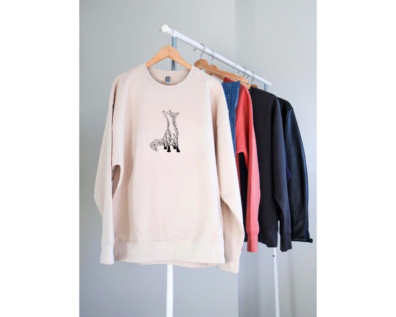 May include: A beige crewneck sweatshirt featuring a black line art illustration of a fox sitting and looking upward. The sweatshirt is displayed on a wooden hanger on a clothing rack alongside other garments in blue, red, black, and dark gray.