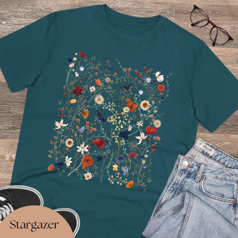 May include: Teal blue t-shirt with a floral print featuring a variety of flowers, butterflies, and leaves in shades of red, pink, yellow, and green.