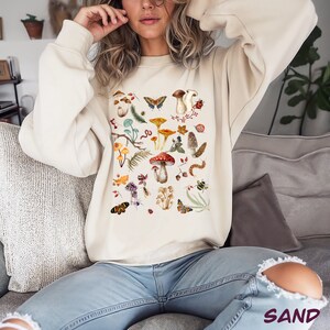 K&ouml;nnte beinhalten: Ein cremefarbener Pullover mit einem bunten Pilz-, Schmetterlings- und Bl&auml;tterdruck. Der Druck zeigt eine Vielzahl von Pilzen, Schmetterlingen und Bl&auml;ttern in Rot-, Orange-, Gelb-, Gr&uuml;n- und Braunt&ouml;nen.