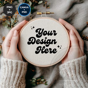 Pode incluir: Uma pessoa está segurando um aro de bordado branco com o texto "Your Design Here" em preto. O aro é cercado por uma moldura de madeira marrom.