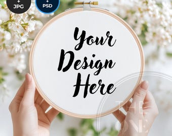 May include: A white embroidery hoop with the text "Your Design Here" in black calligraphy. The hoop is held by hands against a background of white flowers.