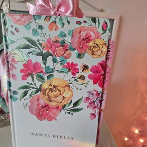 Op de afbeelding: Een boek met een bloemenpatroon en een roze strik. De omslag toont een aquarelpatroon van roze, gele en oranje bloemen op een lichtblauwe achtergrond. De tekst "SANTA BIBLIA" is zichtbaar op de omslag.