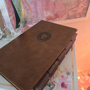 May include: A brown leather-bound journal with a lion emblem. The journal has multiple tabbed sections and is resting on a floral patterned surface. The background includes a framed image of a castle and decorative ribbons.