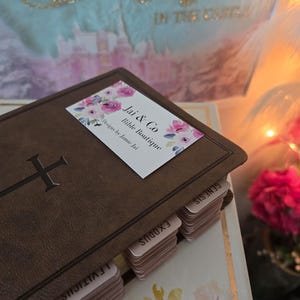 May include: A brown Bible with a cross emblem and labeled tabs. A sticker with floral designs and the text "Jai & Co Bible Boutique" is on the cover. The background includes a blurred image with text and flowers.