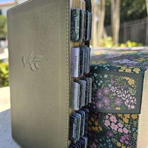 Puede incluir: Biblia verde oliva con páginas doradas y pestañas etiquetadas. La cubierta presenta un diseño de rama de olivo en relieve. Una caja con estampado floral es visible en el fondo. Las pestañas están etiquetadas con nombres de libros.