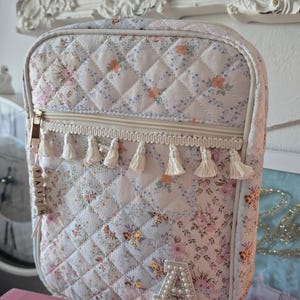 May include: Quilted floral print bag with a zipper closure and white tassels. The bag features a pearl-studded letter "A" and the word "FAITH" on a wooden bead charm. The bag is a soft pastel color with a floral pattern.