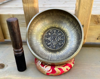 May include: A bronze singing bowl with intricate etched patterns and a central embossed design, resting on a red and gold cushion. A wooden striker with a black felt end sits beside the bowl, suggesting its use for meditation and sound healing.