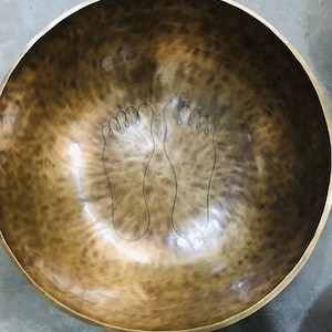 May include: A large, bronze-colored singing bowl with a smooth, reflective surface. The interior features a black outline of two feet. The bowl's rim is a darker shade of bronze. The bowl is likely used for meditation or sound healing.