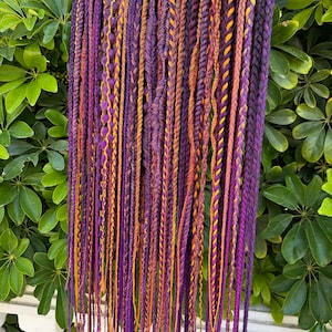 Può includere: Una collezione di extension per capelli intrecciati di colore viola e arancio appese a un appendiabiti. Le extension sono di diverse lunghezze e hanno diverse trame.