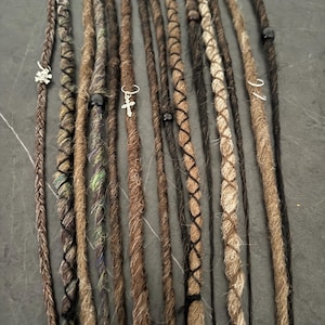 Può includere: Una collezione di dreadlock marroni e beige, alcuni con sezioni intrecciate e ciondoli decorativi. I dreadlock variano per colore e consistenza, con accenti neri e piccoli ciondoli metallici, tra cui un teschio e una croce.