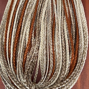 Può includere: Extensioni di capelli sintetici intrecciati in tonalità marrone chiaro, beige e marrone medio. Le trecce sono fittamente intrecciate e disposte in un fascio. I capelli sono legati in basso con un filo abbinato. L'aspetto generale è ordinato e strutturato.