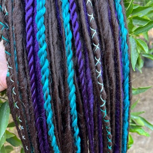 Può includere: Un set di dreadlock sintetiche in marrone, turchese e viola. Le dreadlock sono disposte in una fila verticale e hanno una varietà di trame.