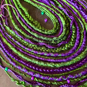 Puede incluir: Primer plano de rastas verdes y moradas. Las rastas están trenzadas y retorcidas, con pequeñas cuentas intercaladas. También se ve una goma para el pelo rosa. El aspecto general es llamativo y colorido.