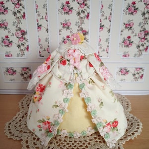 Puede incluir: Un vestido de muñeca de tela amarilla con estampado floral y ribetes verdes y rosas. El vestido está sobre un encaje de ganchillo blanco.