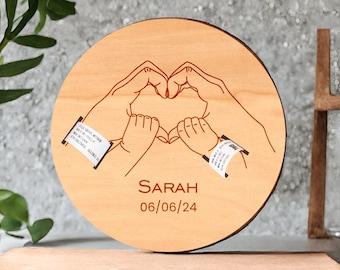 May include: A wooden circle with two hands forming a heart shape. The hands are outlined in red and the circle is light brown. The text "SARAH 06/06/24" is engraved on the circle.