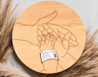 May include: A wooden round plaque with a red outline of three hands holding each other. The hands are of a parent and child. The plaque has a small white tag with the text "Baby Name, Date, Time, Weight, Length" and a QR code.