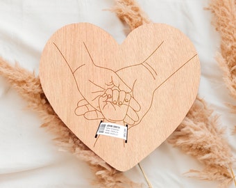 May include: A wooden heart-shaped plaque with a laser-cut design of three hands holding each other. The hands are stylized and represent a family. The plaque is a light brown color and has a natural wood grain.