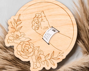 May include: A wooden round sign with a hand reaching out with a wristband that says "Say What A Jella Green" with a barcode. The sign is decorated with a floral design.