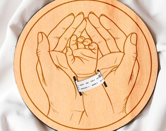 May include: A wooden round plaque with a laser-engraved design of two hands holding a baby's hand. The baby's wrist has a white hospital bracelet with the text "NEWBORN T. SCHNEIDER 8" on it.