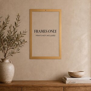 Op de afbeelding: Een rechthoekige, goudkleurige wandlijst met een dunne rand. In het midden van de lijst staat de zwarte tekst "FRAMES ONLY", gevolgd door "PRINTS NOT INCLUDED" in een kleiner lettertype. De lijst is bevestigd aan een effen, neutrale muur.