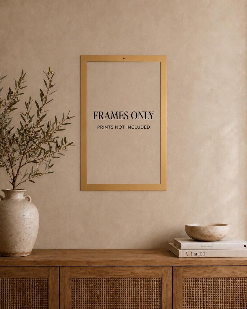 Op de afbeelding: Een rechthoekige, goudkleurige wandlijst met een dunne rand. In het midden van de lijst staat de zwarte tekst "FRAMES ONLY", gevolgd door "PRINTS NOT INCLUDED" in een kleiner lettertype. De lijst is bevestigd aan een effen, neutrale muur.