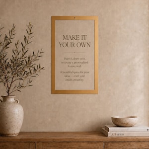Op de afbeelding: Een rechthoekige goudkleurige lijst aan een muur, met een beige bordje met de tekst: MAKE IT YOUR OWN. Paint it, draw on it, or create a personalized feature wall. A beautiful space for your ideas&mdash;even your child's creativity.