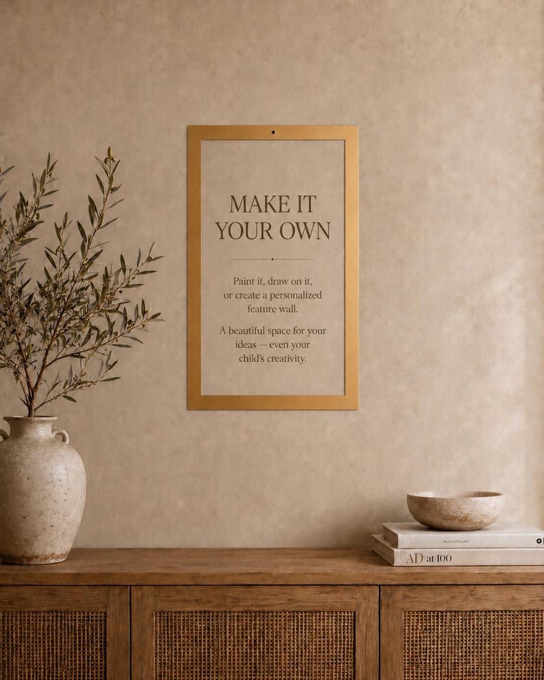Op de afbeelding: Een rechthoekige goudkleurige lijst aan een muur, met een beige bordje met de tekst: MAKE IT YOUR OWN. Paint it, draw on it, or create a personalized feature wall. A beautiful space for your ideas&mdash;even your child's creativity.