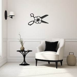 Puede incluir: Arte mural de metal negro que representa unas tijeras con un adorno floral. Las tijeras están abiertas, y la flor está centrada donde se encuentran las hojas. La obra de arte está montada en una pared blanca sobre un sillón blanco.