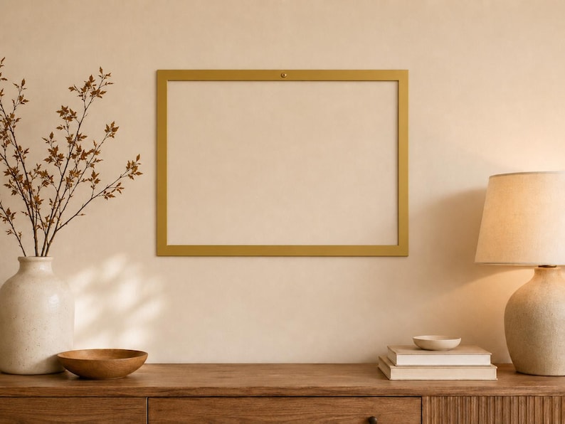 Op de afbeelding: Een rechthoekige goudkleurige fotolijst bevestigd aan een muur. De lijst heeft een dunne, platte rand met een kleine bevestiging zichtbaar in het midden bovenaan. De binnenkant van de lijst is leeg en toont de muur erachter.