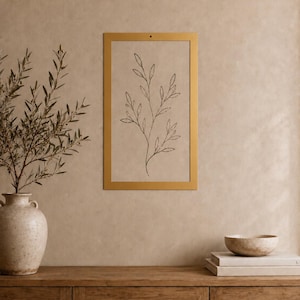 Op de afbeelding: Een rechthoekig wandkunstwerk met een dunne gouden lijst en een beige achtergrond met een zwarte lijntekening van een tak met bladeren. Het kunstwerk hangt aan een muur boven een houten kast met een vaas en een stapel boeken.