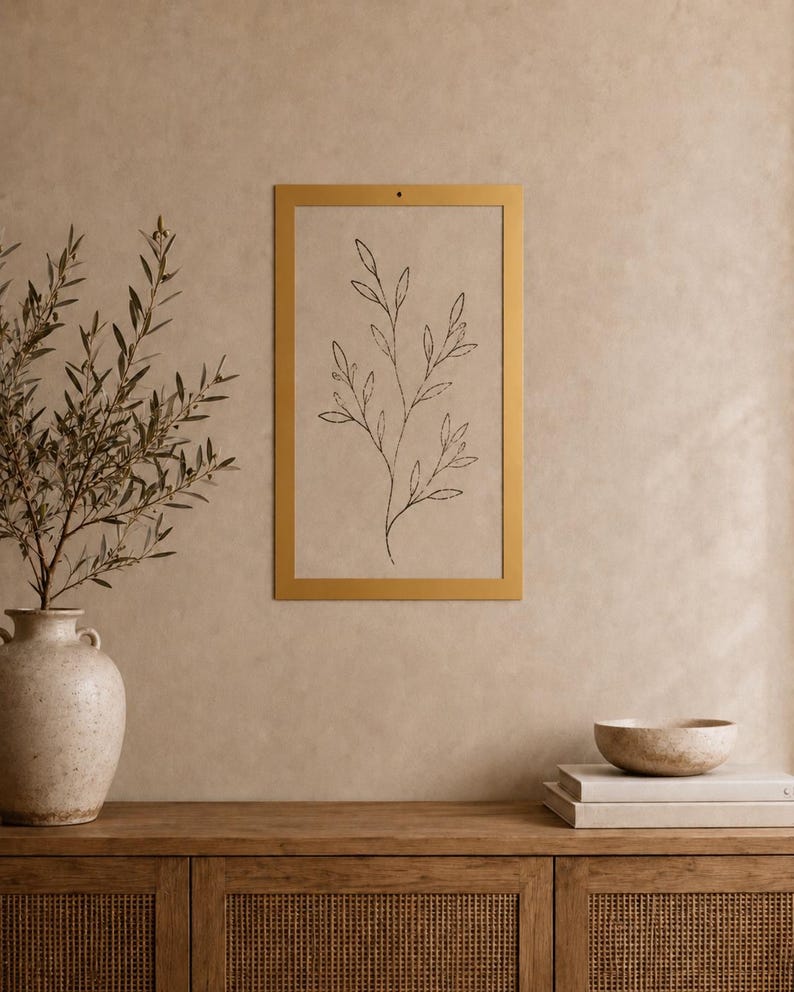 Op de afbeelding: Een rechthoekig wandkunstwerk met een dunne gouden lijst en een beige achtergrond met een zwarte lijntekening van een tak met bladeren. Het kunstwerk hangt aan een muur boven een houten kast met een vaas en een stapel boeken.
