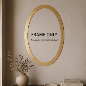 May include: An oval-shaped gold picture frame mounted on a wall. The center of the frame features black text that reads: FRAME ONLY. No paper or insert included. The frame has a smooth, metallic finish and a simple, clean-lined design.