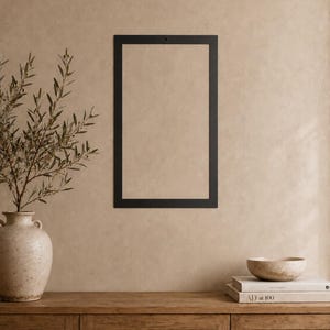 Op de afbeelding: Een rechthoekig zwart metalen frame met een klein gaatje in het midden aan de bovenkant voor bevestiging. Het frame is leeg en heeft een strak, plat en solide randontwerp dat een scherp contrast vormt met de omliggende muur.