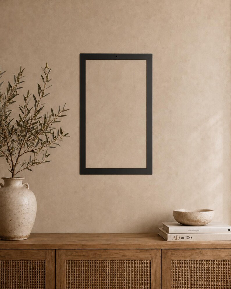 Op de afbeelding: Een rechthoekig zwart metalen frame met een klein gaatje in het midden aan de bovenkant voor bevestiging. Het frame is leeg en heeft een strak, plat en solide randontwerp dat een scherp contrast vormt met de omliggende muur.