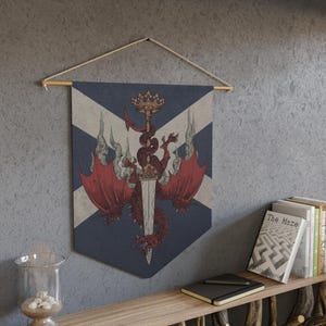 May include: A wall hanging featuring a heraldic design with a red dragon wrapped around a sword, topped with a crown. The background is a blue and white cross design. The banner is suspended from a wooden rod with a natural cord.