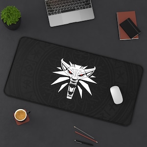 May include: A black gaming mouse pad with a white wolf head design and red eyes. The wolf head is surrounded by a circular pattern. The mouse pad is on a black surface.