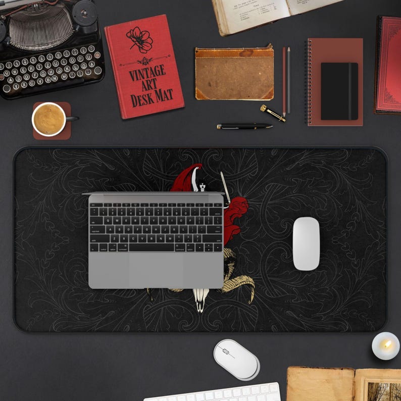 Puede incluir: Una alfombrilla de escritorio rectangular grande con fondo negro, intrincados patrones florales en gris oscuro y una ilustraci&oacute;n gr&aacute;fica central en rojo y dorado de una figura, situada bajo un port&aacute;til plateado y un rat&oacute;n de ordenador blanco.