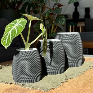 Puede incluir: Tres macetas de cerámica negra con un diseño ondulado y texturado. Las macetas están dispuestas en fila sobre una superficie de madera con una estera de tela verde debajo.