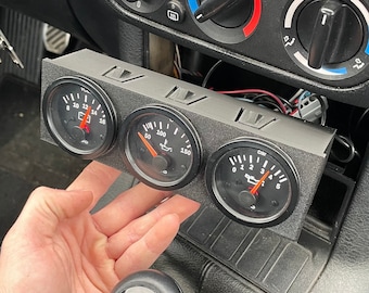 May include: A black gauge pod with three round gauges. The gauges have white faces with black markings and red needles. The gauges are labeled with "Oil", "PSI", and "Bar".
