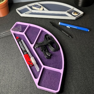 May include: A light purple desk organizer with a purple felt interior, holding pens, a cable, and a clear ruler. A gray organizer with a blue interior is in the background, along with a pencil and eraser. The desk accessories are arranged on a dark gray surface.
