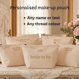 Puede incluir: Bolsas de maquillaje personalizadas beige con texto dorado, incluyendo nombres como "Charlotte" y "Amy bride to be". Las bolsas se exhiben sobre una superficie blanca con un espejo con marco dorado en el fondo. También se ve un ramo de rosas rosas.